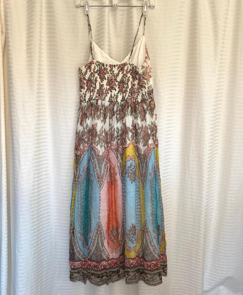 May include: A colorful, patterned maxi dress with spaghetti straps. The dress features a floral bodice and a skirt with vertical panels of blue, pink, yellow, and orange paisley designs. The dress is hanging on a white hanger against a white curtain.