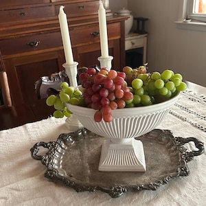 May include: A white ceramic pedestal bowl with vertical ribbed detailing filled with clusters of red and green grapes, resting on an ornate, tarnished silver-toned metal serving tray with decorative handles and a scalloped edge.