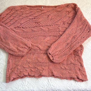 Puede incluir: Un su&eacute;ter de punto color coral con cuello barco y mangas largas. El su&eacute;ter presenta un dise&ntilde;o de punto trenzado texturizado en el cuerpo y las mangas. Las mangas tienen una forma ligeramente abullonada en los hombros y se estrechan hacia las mu&ntilde;ecas.