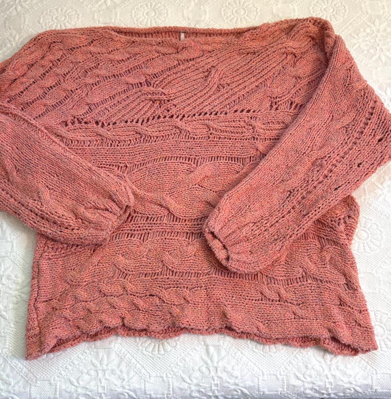 Puede incluir: Un su&eacute;ter de punto color coral con cuello barco y mangas largas. El su&eacute;ter presenta un dise&ntilde;o de punto trenzado texturizado en el cuerpo y las mangas. Las mangas tienen una forma ligeramente abullonada en los hombros y se estrechan hacia las mu&ntilde;ecas.