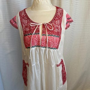 Op de afbeelding: Witte katoenen jurk met rode en roze borduursels. De jurk heeft een ronde hals en korte mouwen. De borduursels zijn in traditionele Mexicaanse stijl.