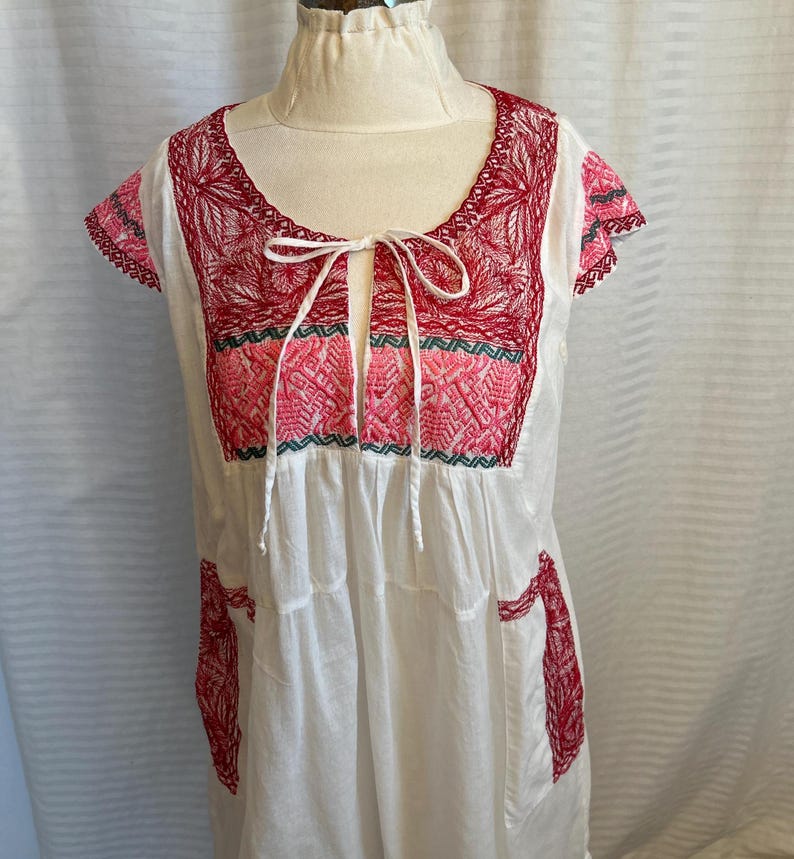Op de afbeelding: Witte katoenen jurk met rode en roze borduursels. De jurk heeft een ronde hals en korte mouwen. De borduursels zijn in traditionele Mexicaanse stijl.