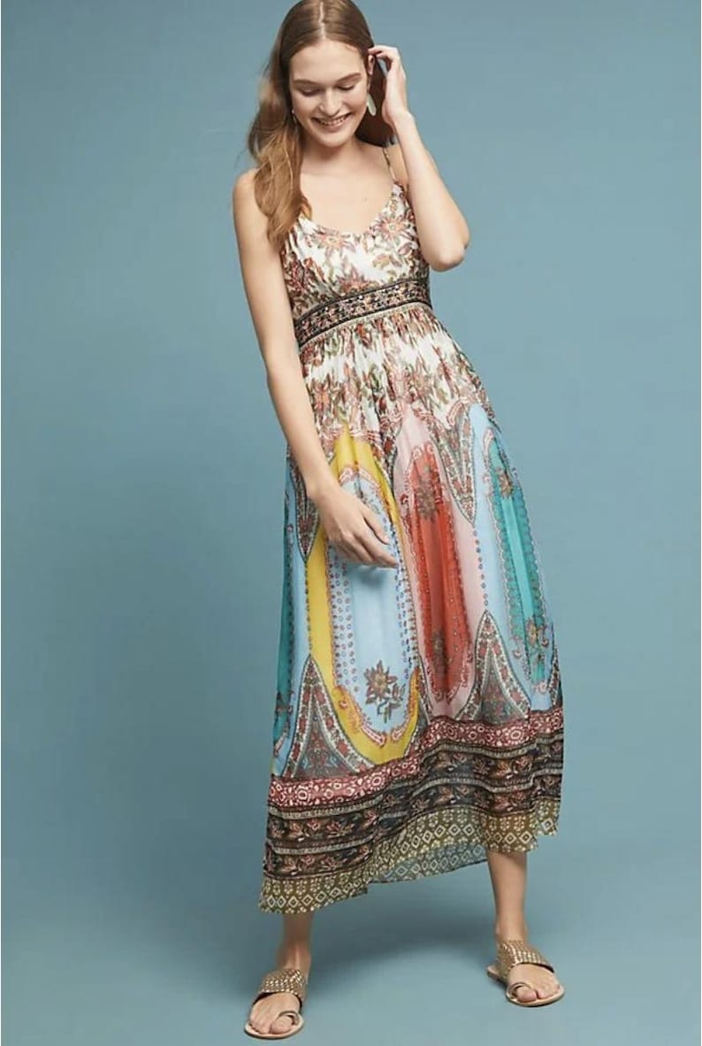 May include: A colorful maxi dress with a floral and paisley print. The dress features a V-neck, spaghetti straps, and a fitted bodice with a patterned waistband. The skirt has vertical panels of yellow, orange, blue, and teal. The dress is worn with gold sandals.
