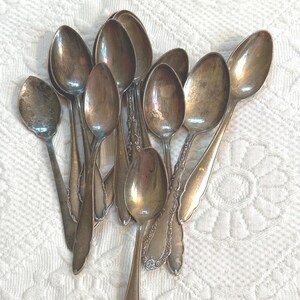 May include: A collection of nine silver-toned metal spoons with varying bowl sizes and handle designs. The spoons exhibit a tarnished, aged patina across their surfaces, with some handles featuring embossed decorative patterns and others being plain.