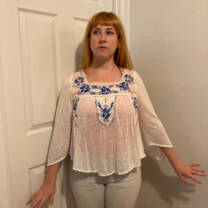 May include: A white, semi-sheer blouse with blue and white embroidery. The blouse has a scoop neck and 3/4 sleeves.