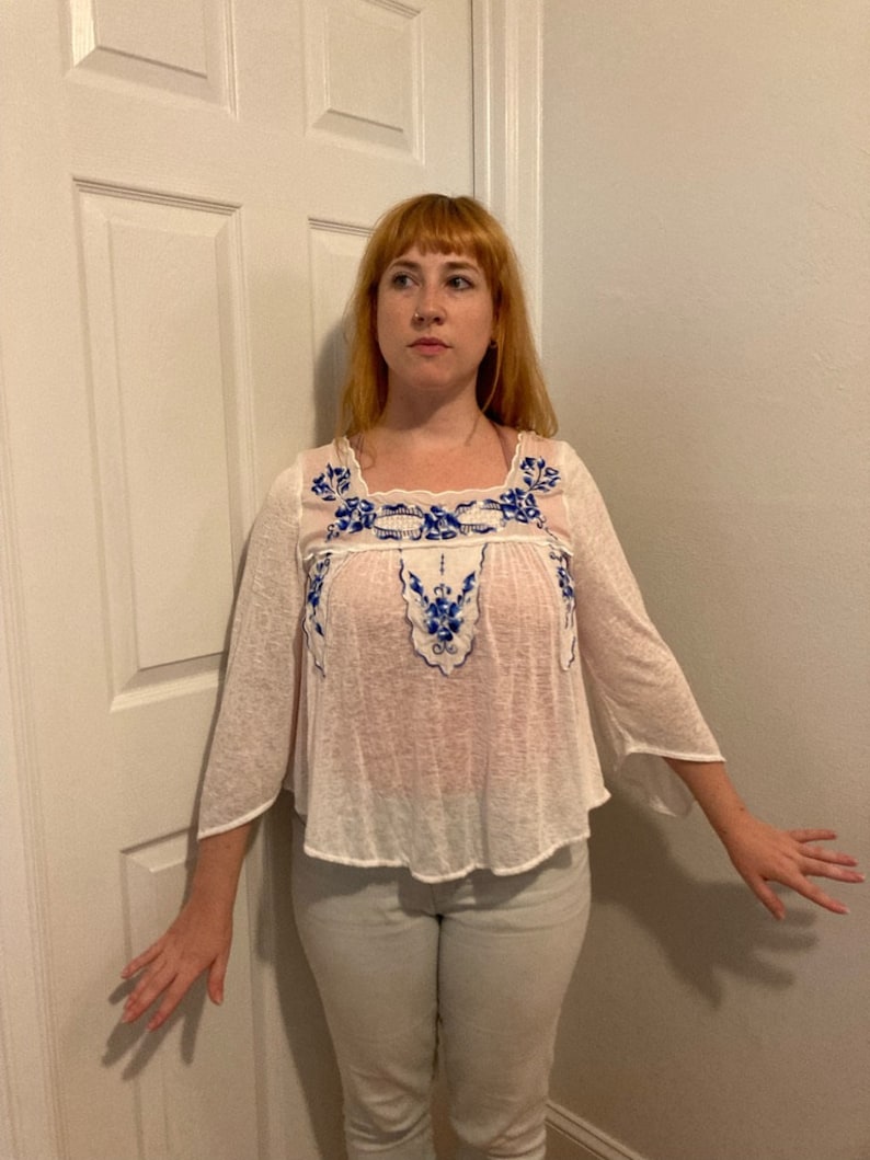 May include: A white, semi-sheer blouse with blue and white embroidery. The blouse has a scoop neck and 3/4 sleeves.
