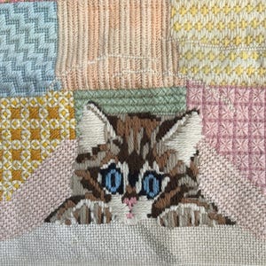 May include: Needlepoint art featuring a brown and white tabby kitten with bright blue eyes. The kitten is set against a patchwork background of various colors and textures, including pink, yellow, and green. The needlepoint is detailed and colorful.