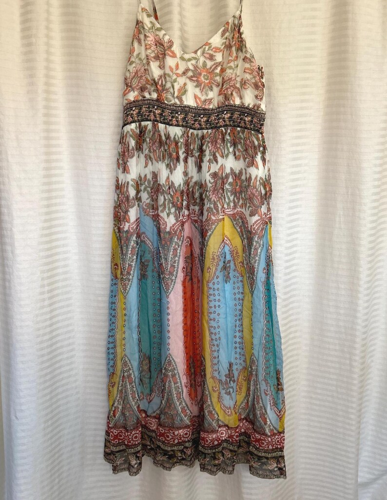 May include: A long, flowing dress with a V-neck and spaghetti straps. The dress features a white bodice with a floral pattern and a colorful, patterned skirt in shades of blue, yellow, orange, and red. The waist has a decorative band.