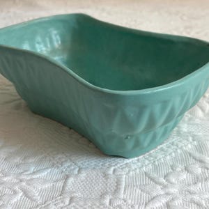 May include: A rectangular turquoise ceramic bowl with rounded corners and a wavy, undulating rim. The exterior walls of the bowl feature a repeating geometric diamond pattern embossed into the surface, providing a textured appearance.