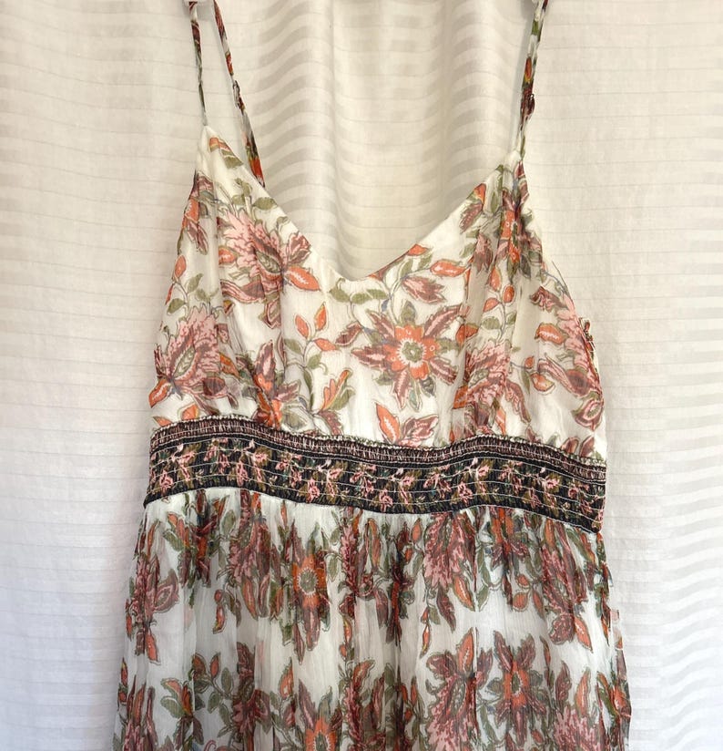 May include: A white sundress with a floral pattern in shades of orange, pink, and green. The dress has thin straps and a fitted bodice with a decorative band. The skirt flows loosely.