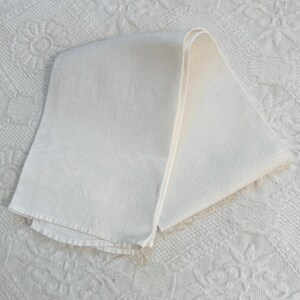 May include: A folded, cream-coloured linen towel. The towel is rectangular with a textured surface. The edges are neatly hemmed. The towel is lying on a white, patterned surface.