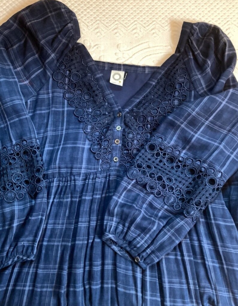 K&ouml;nnte beinhalten: Ein marineblaues kariertes Kleid mit langen &Auml;rmeln. Das Kleid hat einen quadratischen Ausschnitt und ist mit aufwendigen Spitzenverzierungen am Ausschnitt und an den Manschetten versehen. Das Kleid hat eine Knopfleiste vorne und einen flie&szlig;enden, plissierten Rock.
