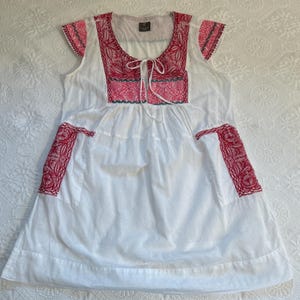 Op de afbeelding: Witte, mouwloze jurk met rode en blauwe borduursels. De jurk heeft een geplooide rok en twee zakken aan de zijkanten.