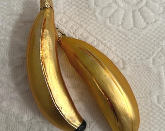 May include: Two gold-coloured banana-shaped Christmas ornaments. Each ornament has a black tip and a gold cap with a wire loop for hanging. The ornaments are on a white, textured surface.
