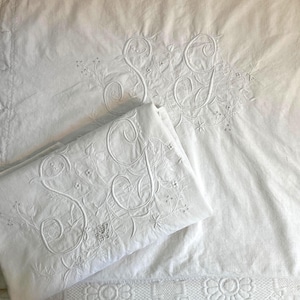 May include: White embroidered bedding set featuring the letters "S" and "J" in a floral design. The set includes a folded pillowcase and a sheet, both with detailed embroidery. The bedding has a vintage aesthetic.