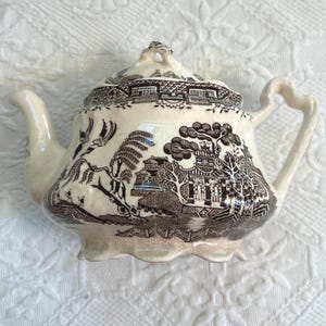 May include: A cream-colored ceramic teapot featuring a dark brown transferware pattern of a traditional willow landscape with pagodas, trees, and birds. The teapot has a scalloped base, a curved handle, a spout, and a matching lid with a finial.