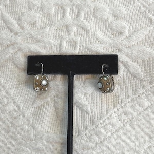 May include: A pair of square earrings with a silver frame and a gold centre. Each earring has a small, round, white stone in the middle, surrounded by small, silver dots. The earrings are displayed on a black stand.