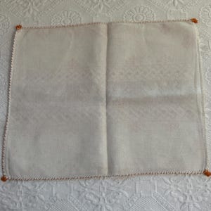 Puede incluir: Un pa&ntilde;uelo de lino blanco con un borde bordado de color naranja. El pa&ntilde;uelo est&aacute; doblado por la mitad y tiene una superficie ligeramente texturizada.