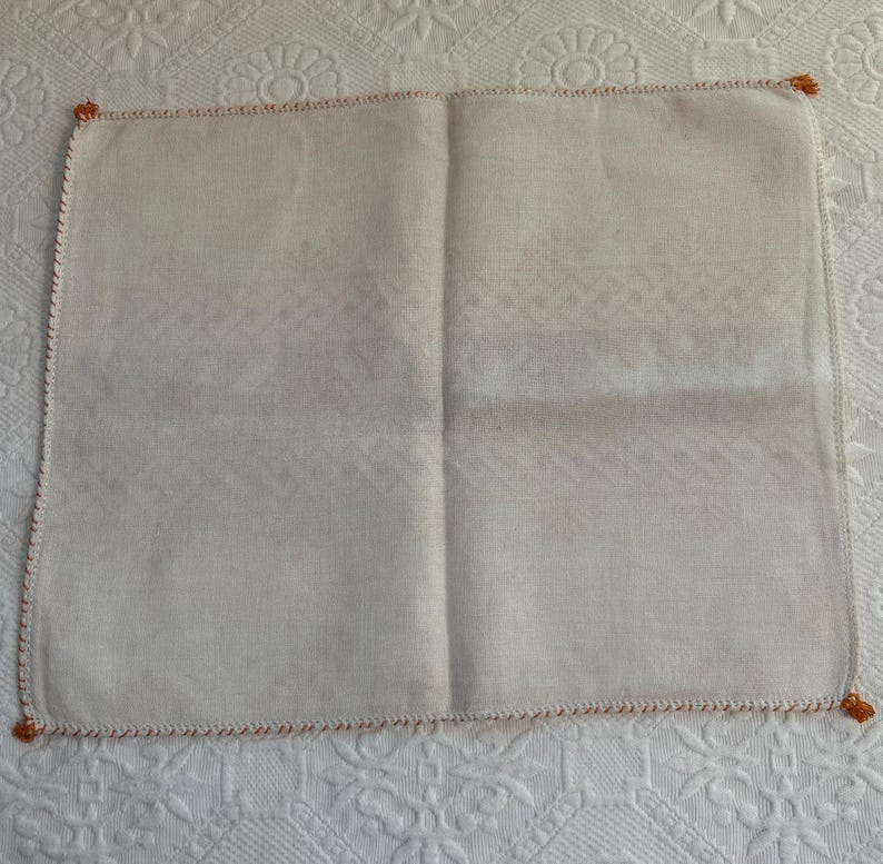 Puede incluir: Un pa&ntilde;uelo de lino blanco con un borde bordado de color naranja. El pa&ntilde;uelo est&aacute; doblado por la mitad y tiene una superficie ligeramente texturizada.