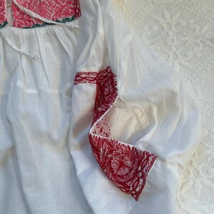 Op de afbeelding: Witte katoenen blouse met rode en groene borduursels rond de halslijn en mouwen. De borduursels zijn een bloemenpatroon.