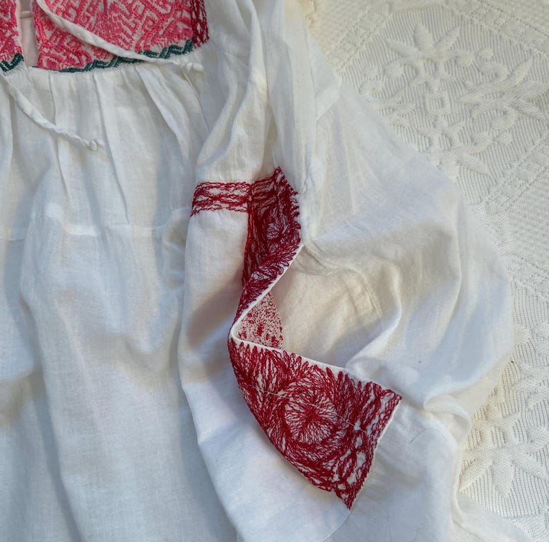 Op de afbeelding: Witte katoenen blouse met rode en groene borduursels rond de halslijn en mouwen. De borduursels zijn een bloemenpatroon.