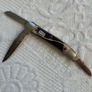 Könnte beinhalten: Ein Taschenmesser mit zwei Klingen und einem braunen und schwarzen Griff. Das Messer hat einen silbernen Metallrahmen und ein kleines silbernes Metallemblem am Griff.