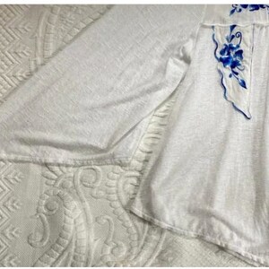 May include: White long-sleeved blouse with blue floral embroidery. The embroidery is a delicate design with blue flowers and leaves.