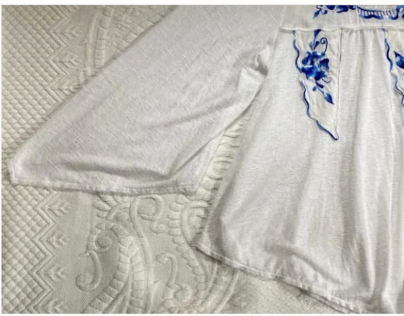 May include: White long-sleeved blouse with blue floral embroidery. The embroidery is a delicate design with blue flowers and leaves.