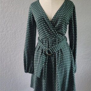 May include: A dark green wrap dress with a white dotted pattern. The dress has long sleeves, a V-neck, and a matching belt with a circular buckle. The dress is displayed on a mannequin.