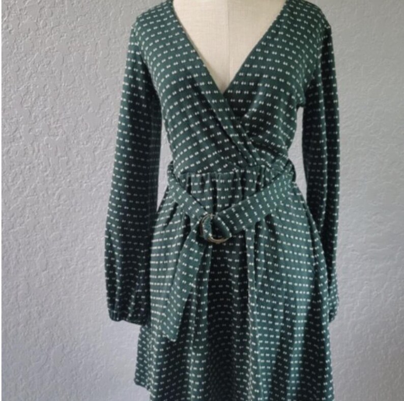 May include: A dark green wrap dress with a white dotted pattern. The dress has long sleeves, a V-neck, and a matching belt with a circular buckle. The dress is displayed on a mannequin.