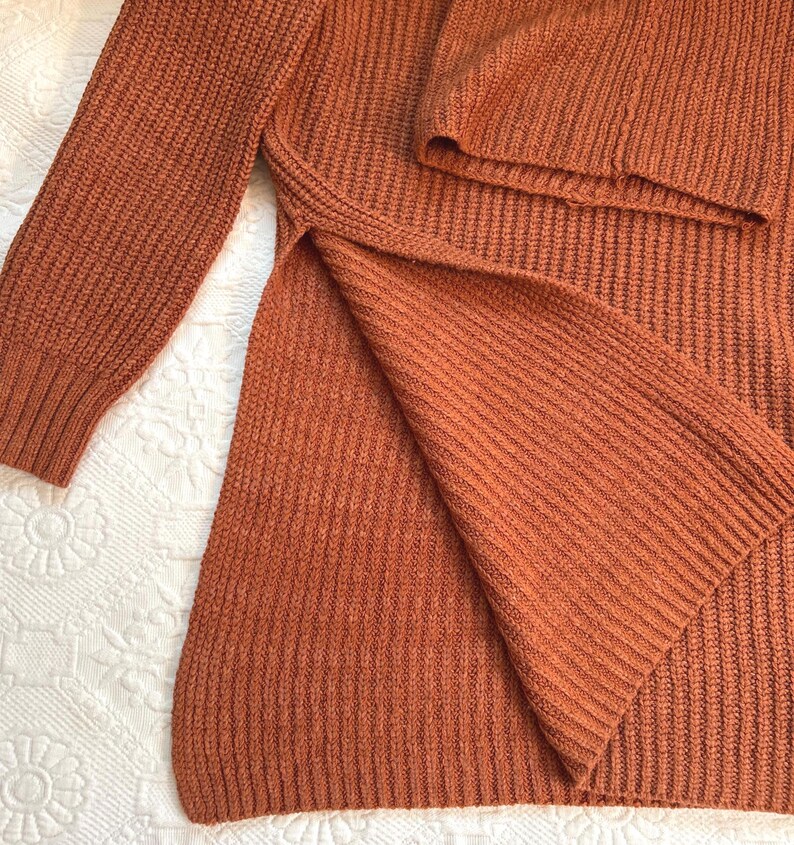 May include: Rust-colored knit sweater with a split hem. The long-sleeved sweater has a ribbed texture and a relaxed fit. The garment is made of a soft, warm material, perfect for cooler weather.