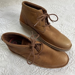 May include: A pair of brown leather ankle boots with laces. The boots have a textured leather upper and a light brown sole. The laces are a matching brown colour. The boots are designed for casual wear.