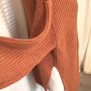 May include: A rust-colored knit sweater with a ribbed texture. The sweater has long sleeves and a split hem on the side. The fabric appears to be a thick, warm material.