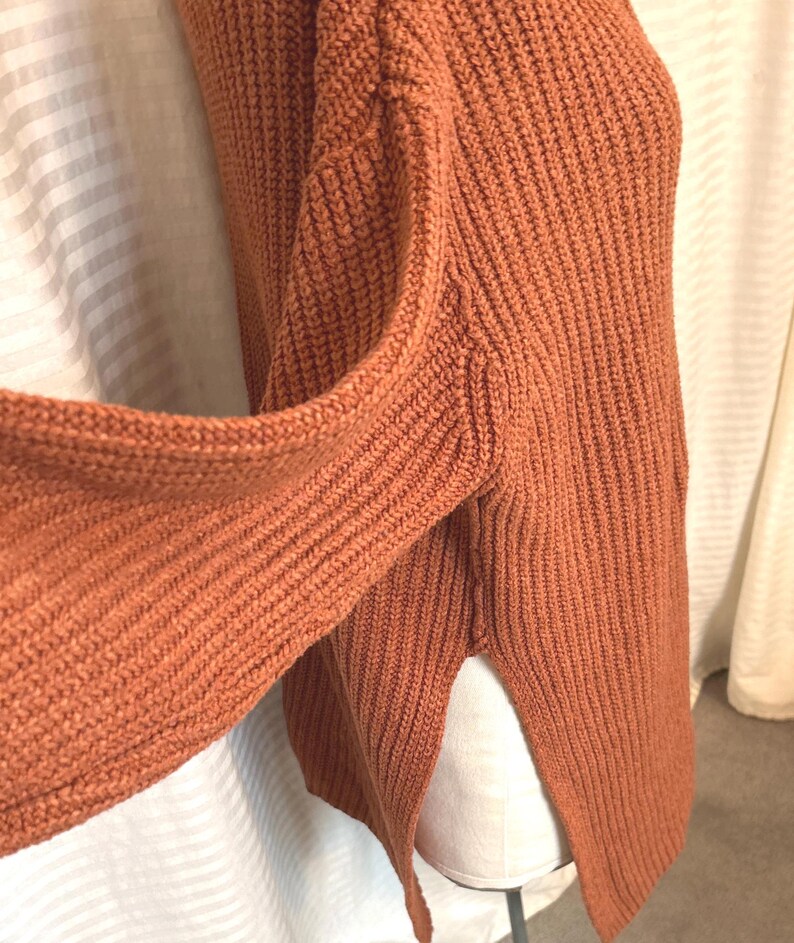 May include: A rust-colored knit sweater with a ribbed texture. The sweater has long sleeves and a split hem on the side. The fabric appears to be a thick, warm material.