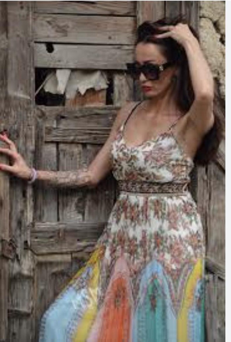 May include: A floral print maxi dress with spaghetti straps. The dress features a white background with a pattern of red and orange flowers. The skirt has a colorful design with blue, orange, and yellow sections. The dress is worn with black sunglasses.