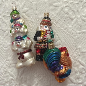 May include: Four glass Christmas ornaments. One is a stack of snowmen, another is a nutcracker, and the last is a colorful rooster. Each ornament has a gold-colored metal cap for hanging.