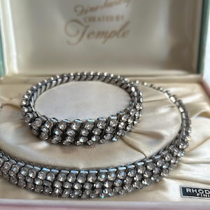 Può includere: Un set di gioielli vintage con bracciale e collana di strass. I pezzi color argento sono esposti in una scatola rosa e crema. La scatola è etichettata "Fine Jewelry Created by Temple".