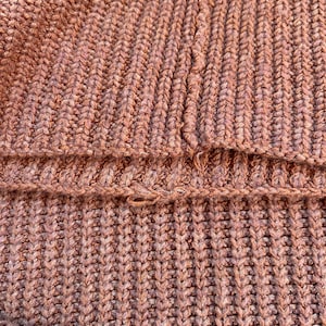 May include: Close-up of a textured, knitted fabric in a muted, dusty rose color. The knit pattern is a thick rib stitch, creating vertical lines. The fabric appears to be a sweater or similar garment, with a visible seam.