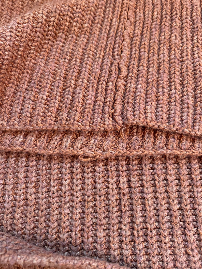 May include: Close-up of a textured, knitted fabric in a muted, dusty rose color. The knit pattern is a thick rib stitch, creating vertical lines. The fabric appears to be a sweater or similar garment, with a visible seam.