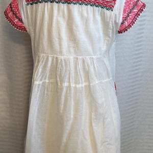Op de afbeelding: Witte katoenen jurk met rode en roze borduursels rond de halslijn en mouwen. De jurk heeft een geplooide rok en een eenvoudig, klassiek ontwerp.