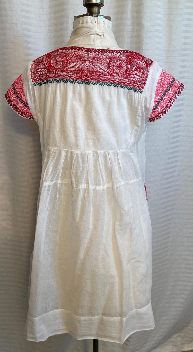 Op de afbeelding: Witte katoenen jurk met rode en roze borduursels rond de halslijn en mouwen. De jurk heeft een geplooide rok en een eenvoudig, klassiek ontwerp.