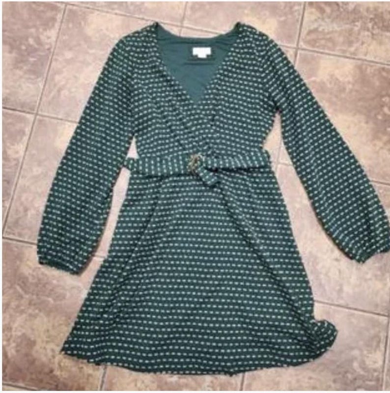 May include: A dark green dress with a white dotted pattern, long sleeves, and a V-neck. The dress features a matching belt with a gold-coloured buckle. The dress is knee-length and made of a lightweight fabric.