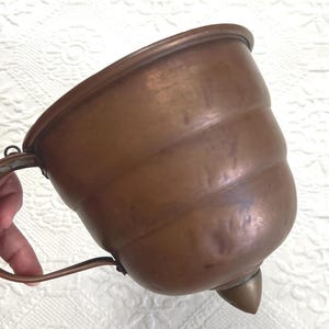 May include: A copper vessel with a rounded, ribbed body and a wide, open top. The container features a curved metal handle attached to the side and a pointed, conical base at the bottom, showcasing a warm, metallic brown finish throughout.