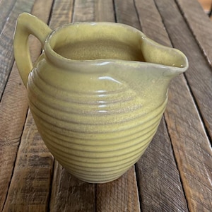 Puede incluir: Una jarra de cerámica amarilla con un cuerpo redondeado con detalles acanalados horizontales, un asa curva en el lateral y un pico vertedor prominente. El esmalte tiene una textura sutil y moteada con variaciones en el color amarillo.