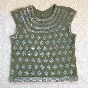 Puede incluir: Un suéter verde sin mangas con lentejuelas plateadas. El suéter tiene un cuello redondo y un patrón de diamantes de lentejuelas en la parte delantera. El cuello y el dobladillo están adornados con lentejuelas plateadas.