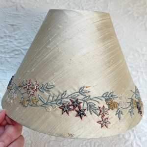 K&ouml;nnte beinhalten: Cremefarbener Lampenschirm mit floralem Stickdesign. Die Stickerei zeigt rote, blaue und goldfarbene sternf&ouml;rmige Blumen und Bl&auml;tter. Der Lampenschirm hat eine leicht konische Form.