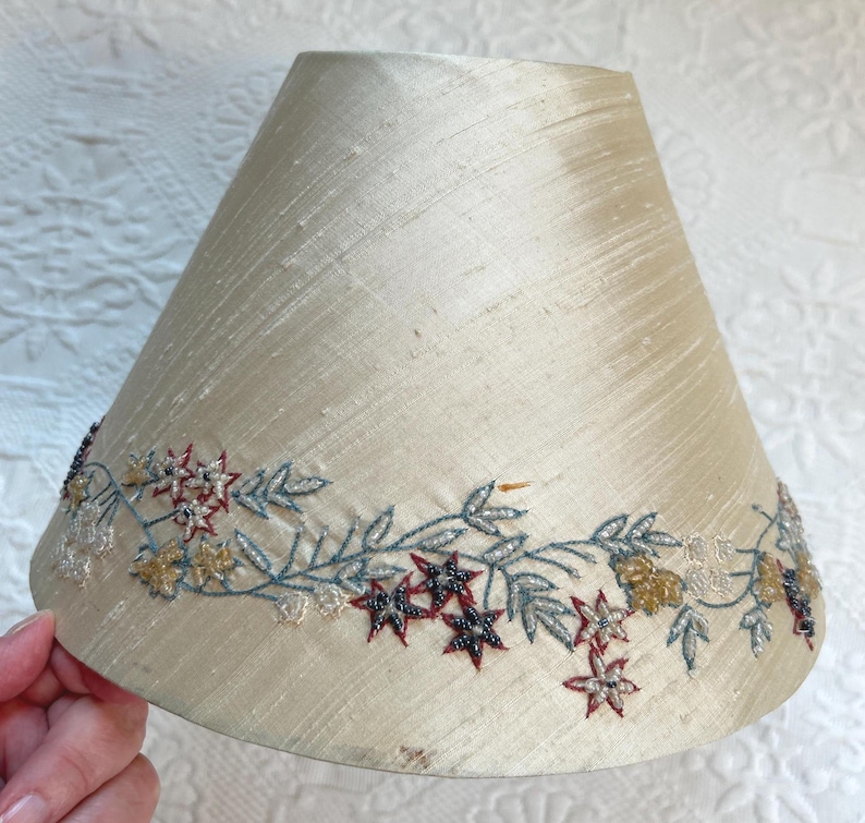 K&ouml;nnte beinhalten: Cremefarbener Lampenschirm mit floralem Stickdesign. Die Stickerei zeigt rote, blaue und goldfarbene sternf&ouml;rmige Blumen und Bl&auml;tter. Der Lampenschirm hat eine leicht konische Form.