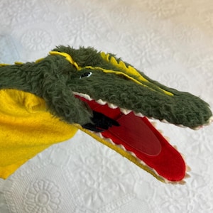 May include: A plush alligator puppet with a green, textured body, yellow accents, and a red mouth. The puppet has white teeth and a black tongue. The puppet is made of soft, fuzzy material.