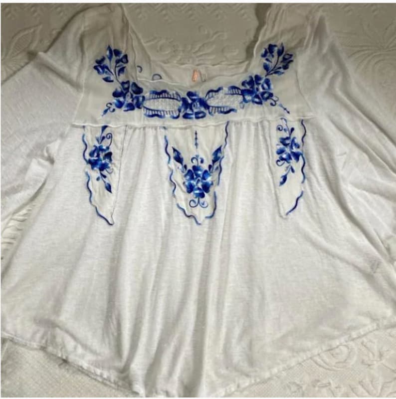 May include: White embroidered blouse with blue floral designs. The blouse has a scoop neck and 3/4 sleeves.
