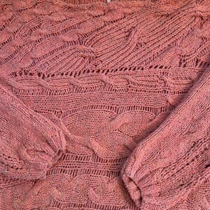 Puede incluir: Un su&eacute;ter de color rosa empolvado con un dise&ntilde;o de punto texturizado. El su&eacute;ter presenta un cuello barco y mangas largas con un patr&oacute;n de punto trenzado. La prenda de punto tiene un aspecto suave y ligeramente descolorido.